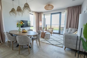 A modern living room with a round dining table, grey sofas, and a large window with curtains.