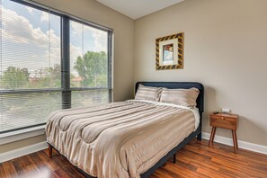 A bedroom with a large window, a bed with a headboard, a bedside table, and a wall-mounted picture frame. - Walkable Condo in Dtwn Greensboro! Extended Stay (Greensboro)