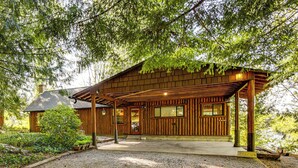 A wooden cabin with a porch, surrounded by trees and a gravel driveway. - Beach Access Historic Cabin on Henderson Bay! (Gig Harbor)