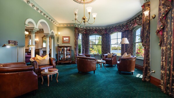 A spacious room with leather armchairs, a chandelier, a fireplace, and large windows with curtains.
