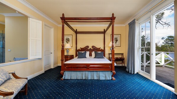 A four-poster bed with a wooden frame, a blue carpet with a subtle pattern, a nightstand with a lamp, and a view of the outdoors through a large window.