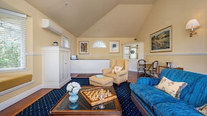 Un salon avec un canapé bleu, une table basse sur laquelle est posé un jeu d’échecs, un meuble blanc et un coin repas avec des chaises en osier.
