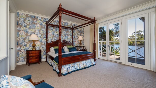 A four-poster bed with a canopy, a wooden nightstand with a lamp, and a view of a body of water through large sliding glass doors.