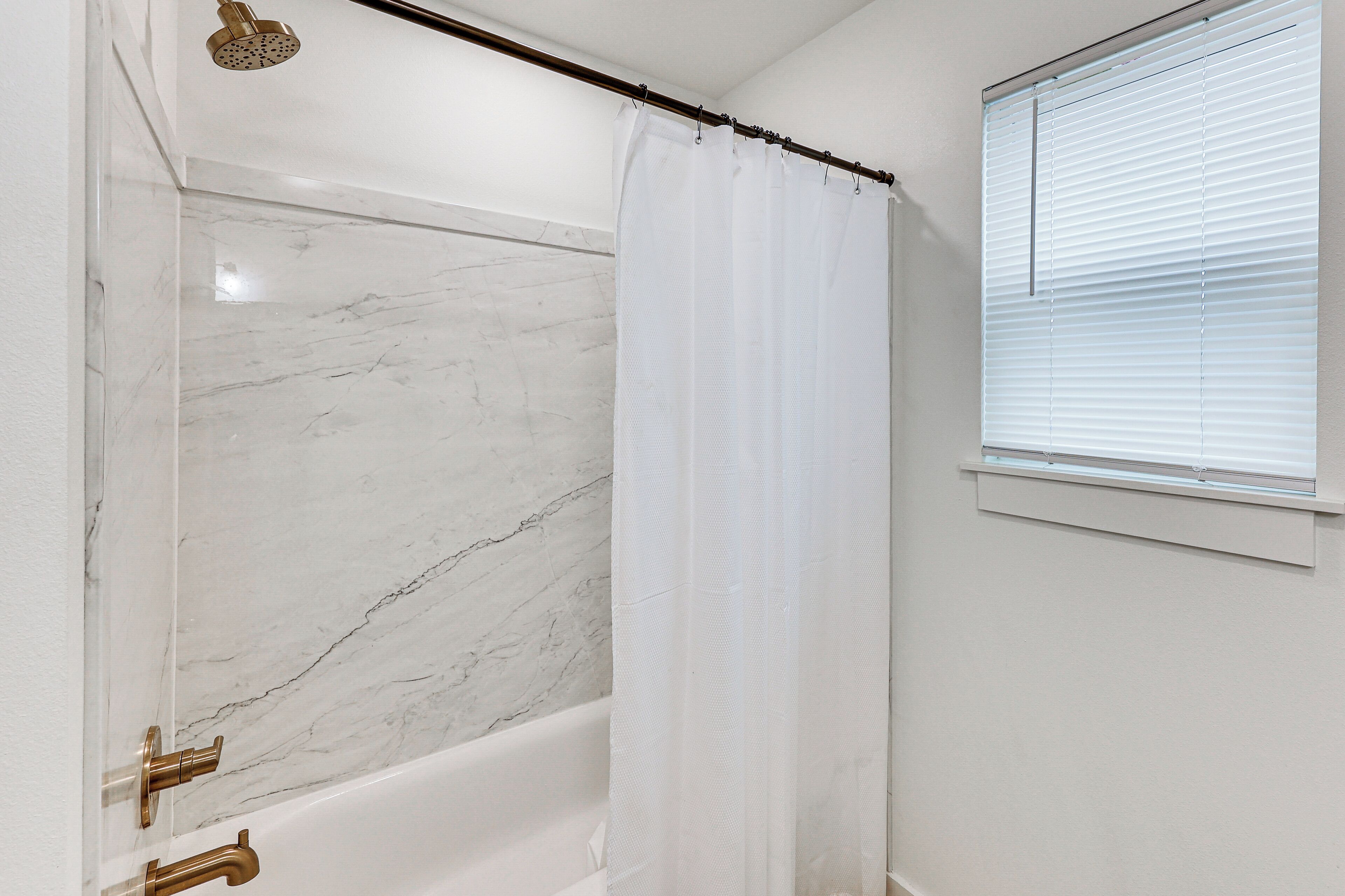 Une salle de bain avec un mur de douche en marbre, une baignoire et une fenêtre avec des stores.