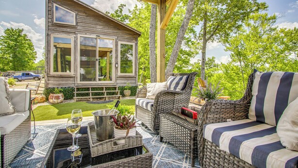 A covered patio with wicker furniture, a table with a wine glass, and a view of a wooden house. - Tiny Cabin Getaway in the Tennessee River Valley (Rogersville)