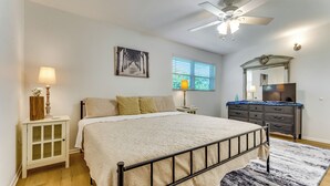 Une chambre à coucher comprenant un lit, une table de chevet, une commode, un ventilateur de plafond, une fenêtre avec des stores et un tableau accroché au mur.