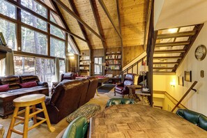 A spacious living area with a high ceiling, wooden beams, a large window overlooking a forest, a fireplace, and a staircase. - Walk to Lake Arrowhead! All-Season Alpine Escape (Lake Arrowhead)