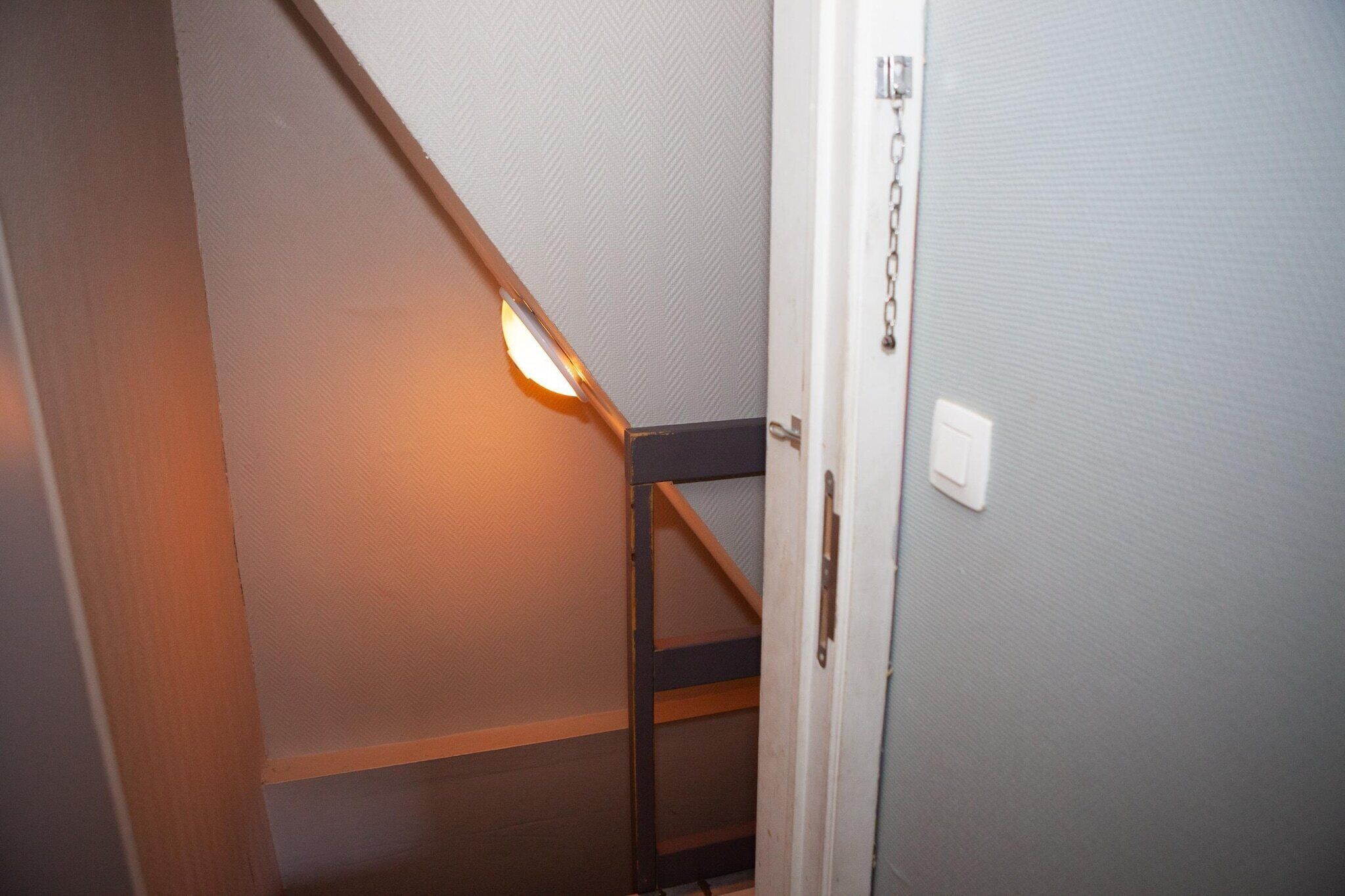 A narrow hallway with a wooden staircase, a door with a chain lock, and a light fixture.