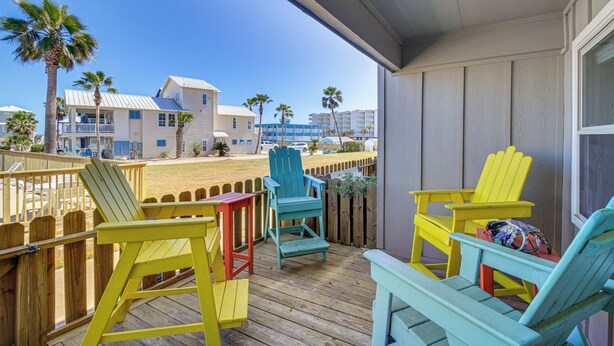 Una terraza de madera con sillas Adirondack de colores, vista a la playa y un edificio con fachada clara.