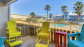 Una terraza de madera con sillas Adirondack de colores, una piscina y vistas a un área residencial con palmeras y casas.