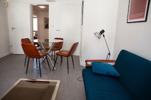 Une pièce comprenant une table à manger et des chaises, un canapé avec un coussin, un lampadaire et un tableau encadré accroché au mur.