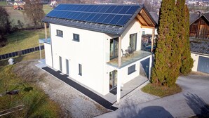 Een modern huis met een dak voorzien van zonnepanelen, een glazen balkon en een hoge conifeer.