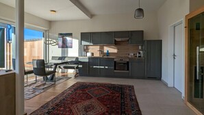 Een moderne keuken met donkere kasten, een eethoek met tafel en stoelen, en een groot raam dat voor natuurlijk licht zorgt.