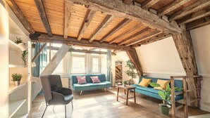 Een zolderkamer met zichtbare houten balken, een blauwe bank, een grijze fauteuil en een houten trap.