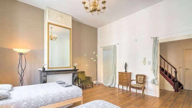 A bedroom with two beds, a large mirror, a chandelier, a staircase, and a green armchair. - Elegant Duplex for 12 in Capital Street (Antwerp)