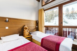 Ein Hotelzimmer mit zwei Betten, einem Holz-Kopfteil, einem Fenster mit Blick auf schneebedeckte Bäume und einem Teddybären auf dem Nachttisch.