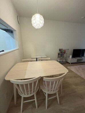 Un coin repas avec une table en bois clair et des chaises blanches, un luminaire suspendu et un téléviseur en arrière-plan.