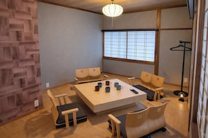 Quarto com piso de madeira, mesa central com cadeiras, luminária pendente e uma grande janela com vista.