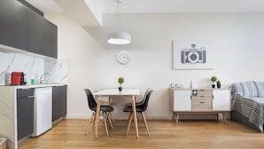Ett modernt kök med en inbyggd diskmaskin, en matplats med ett vitt bord och svarta stolar, ett litet sidobord med en växt och en säng med ett randigt överkast.