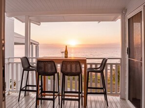 A wooden table with chairs on a balcony overlooking the ocean at sunset.