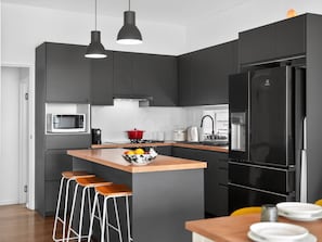 A modern kitchen with dark cabinets, a black refrigerator, a microwave, a red pot, and a fruit bowl on the island.