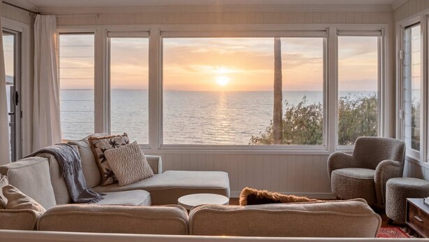 A room with a view of the sunset over the sea, a ceiling fan, and a sofa with a throw blanket.