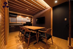A modern kitchen with wooden cabinetry, a dining area with a wooden table and chairs, and a black wall with a mounted television. - Hanaikada House 8minute walk from Kyoto Station / Kyoto City Kyōto (Kyoto City)