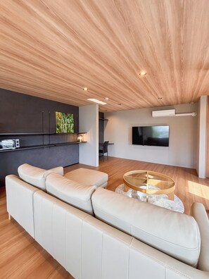 Un salon moderne avec un grand canapé beige, une table basse en verre, un téléviseur à écran plat fixé au mur et un plafond en bois.