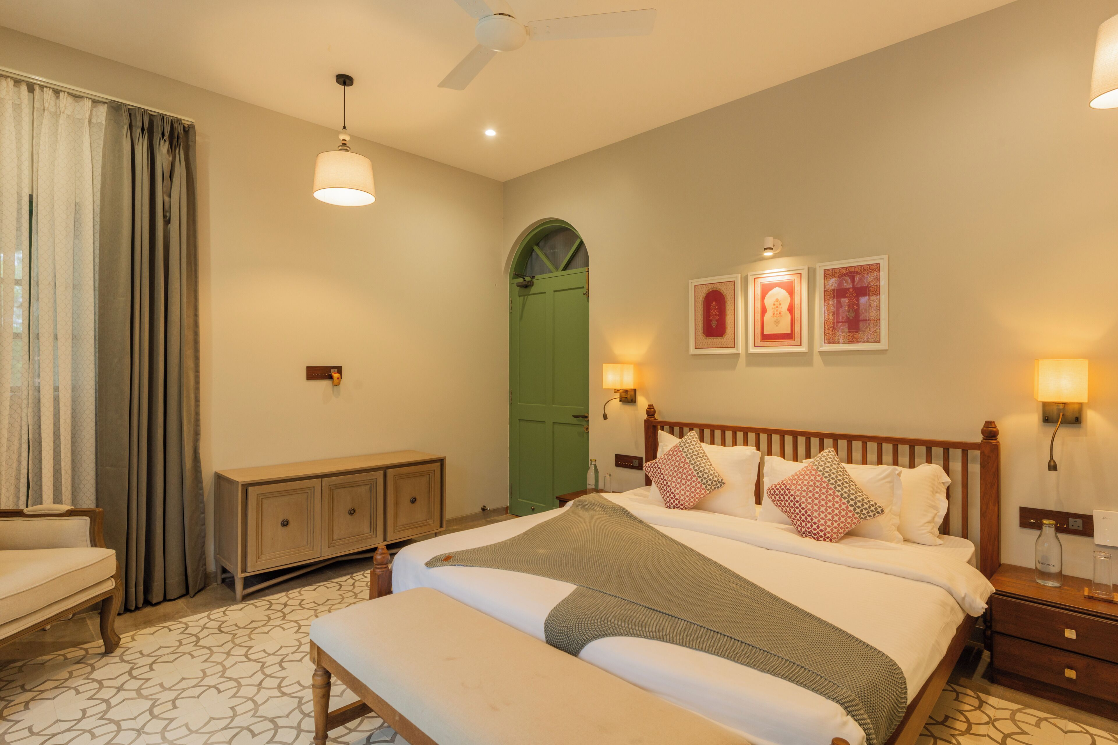 A hotel room with a bed, bedside tables, a bench, a green door, and wall-mounted lights.