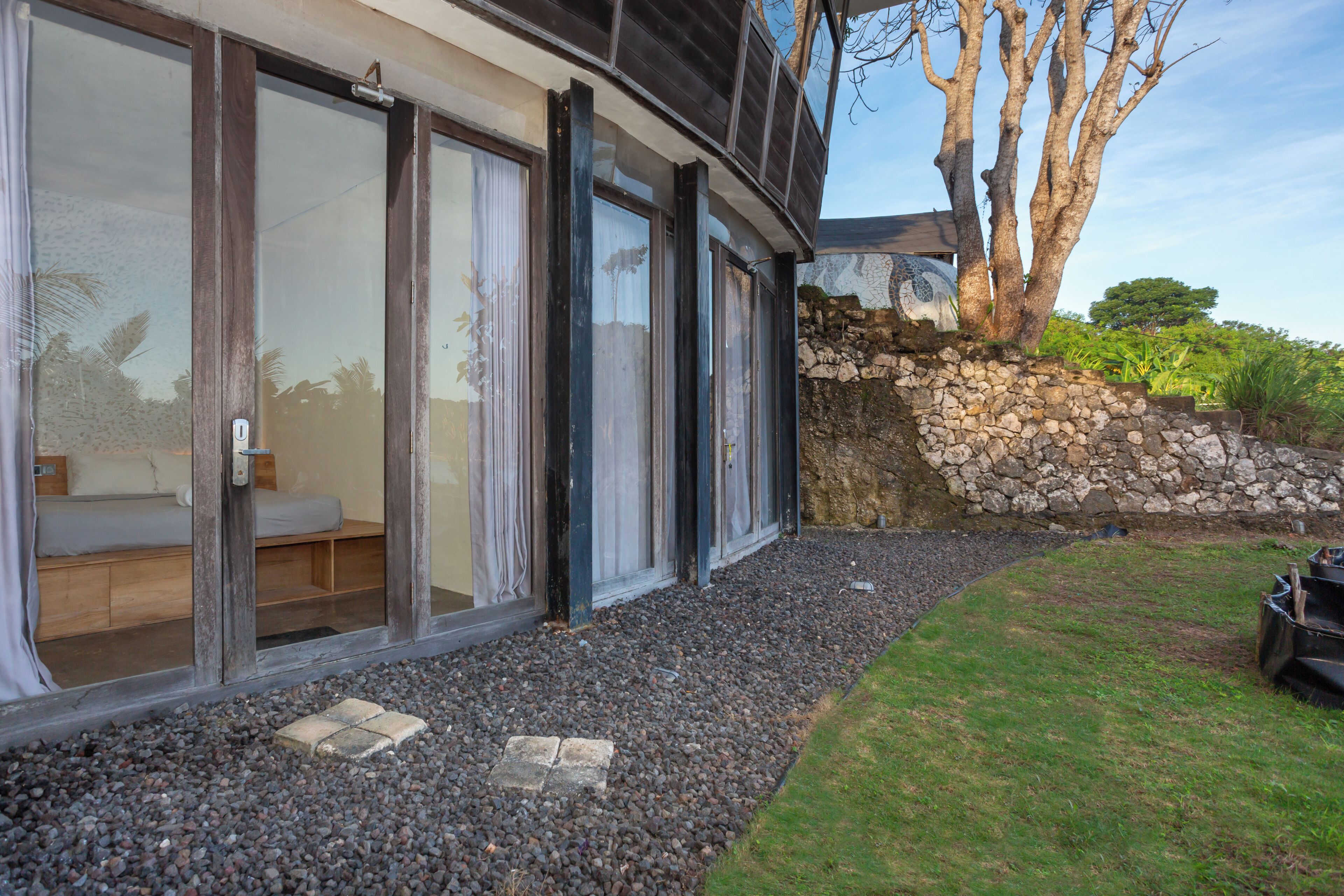 A modern building with large sliding glass doors, a stone wall, and a grassy area with a tree.