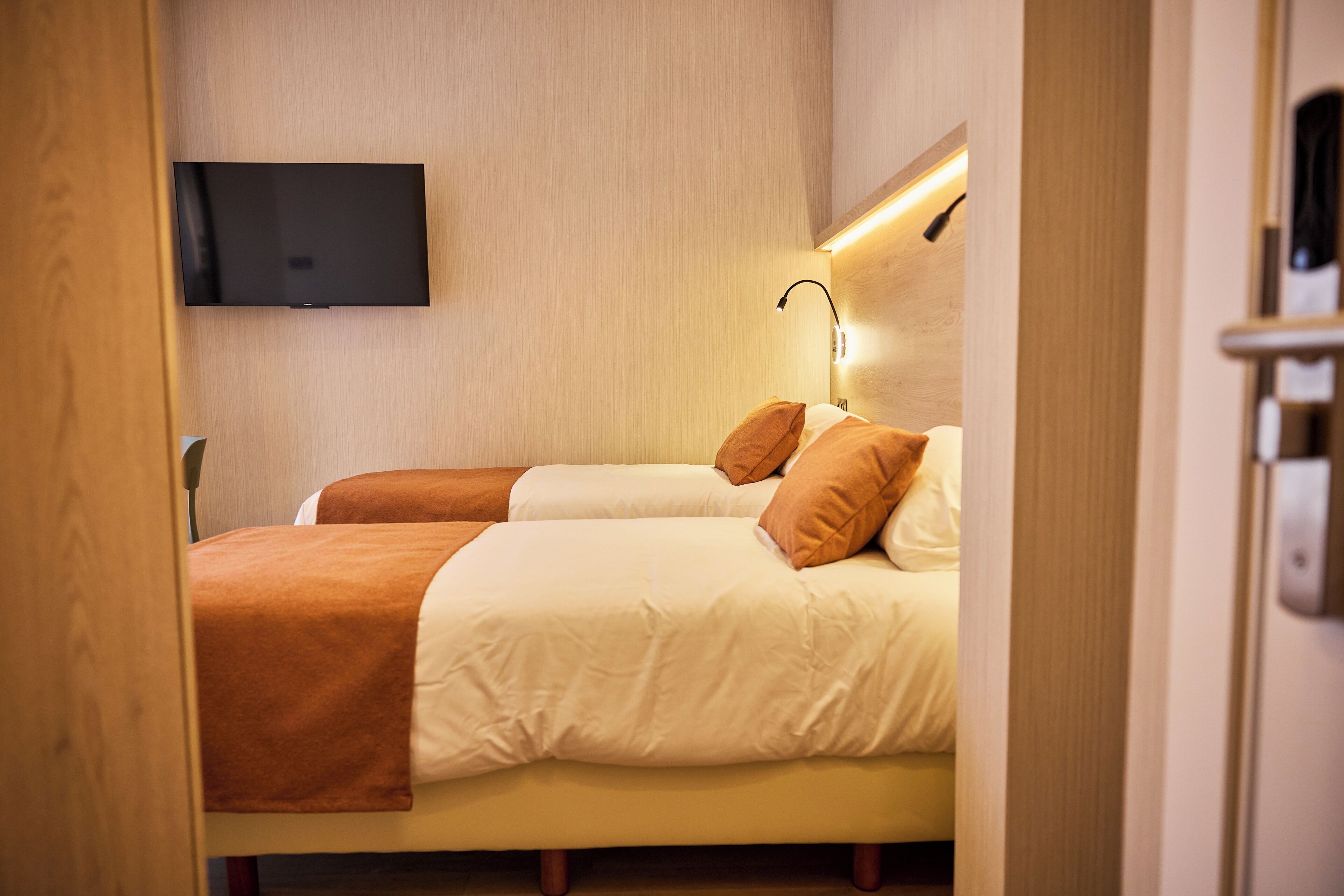 Habitación de hotel con dos camas, un televisor de pantalla plana y una lámpara fijada a la pared.