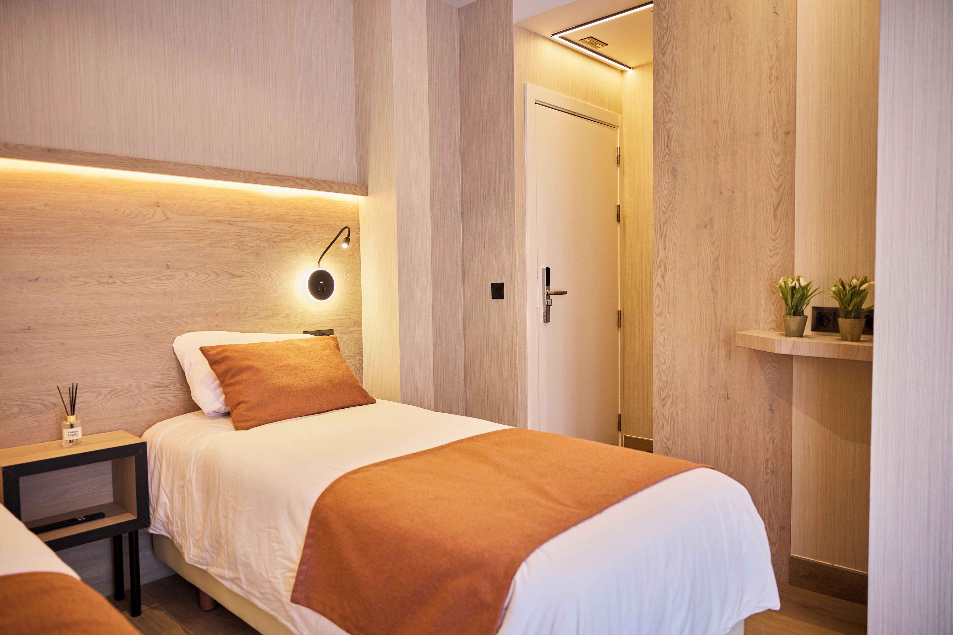 A hotel room with a bed, bedside table, nightstand, and a wall-mounted lamp.
