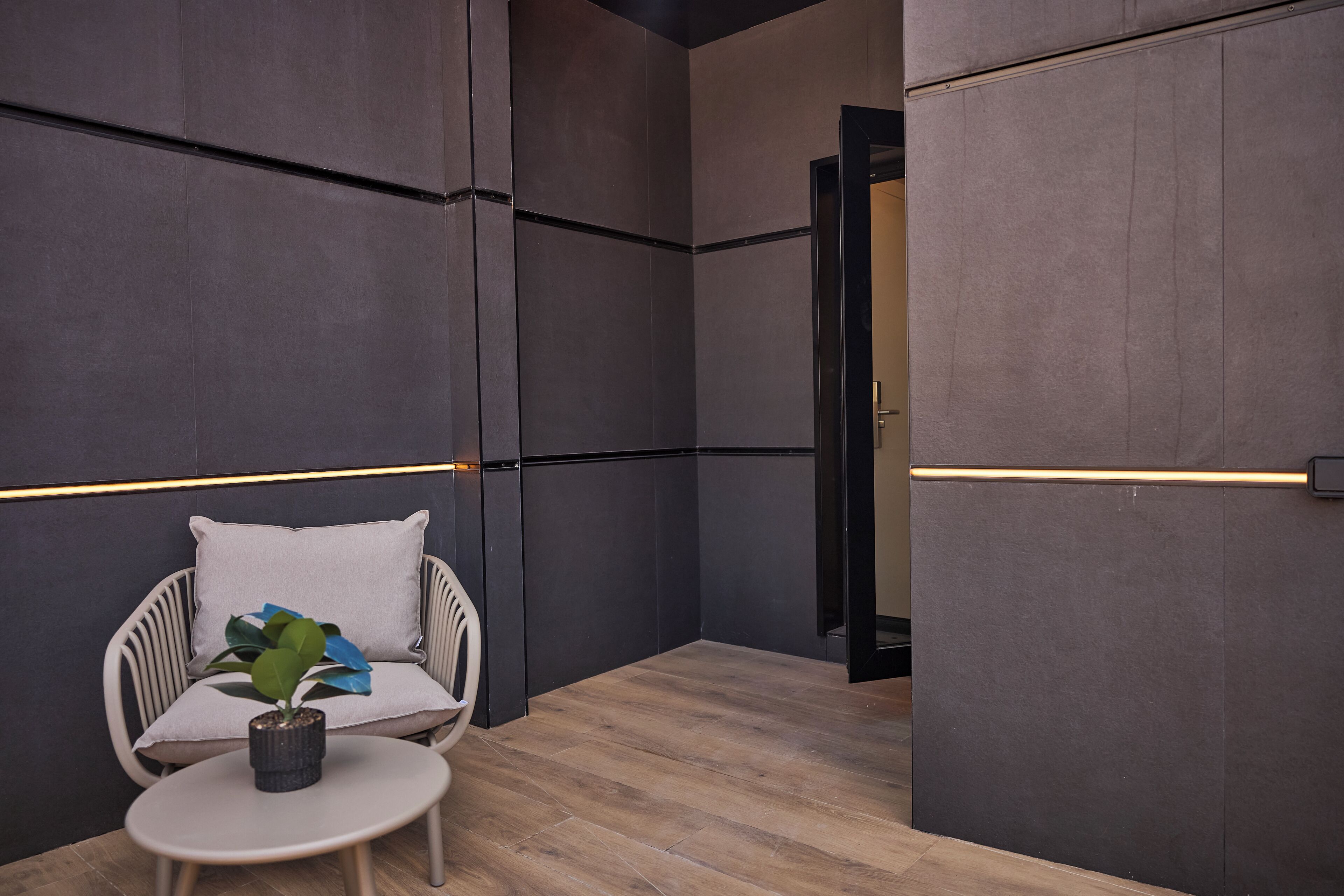 Een moderne kamer met houten vloer, een witte stoel met kussen, een tafeltje met plant en donkere muren met lichtbalk.