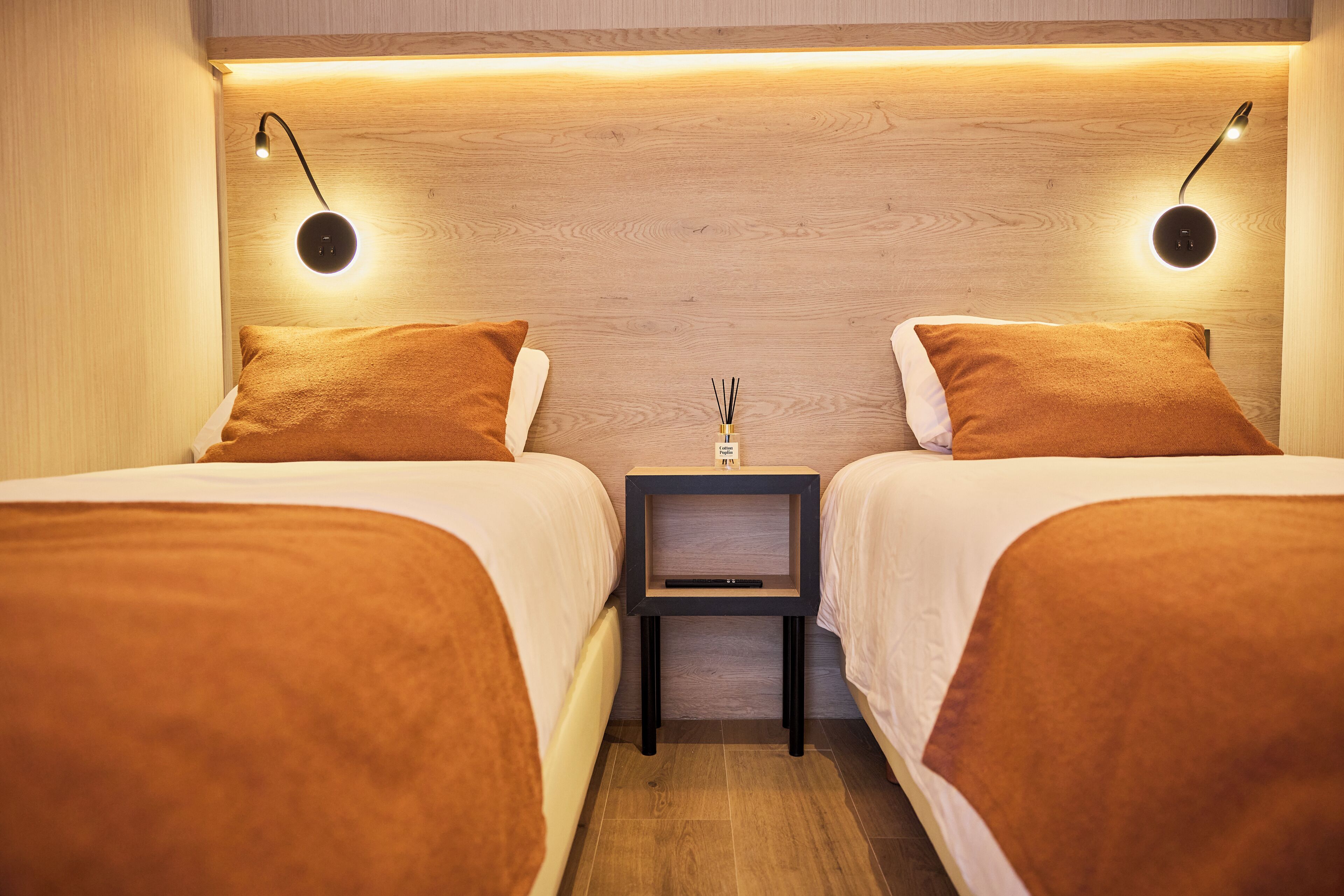 Une chambre avec deux lits, chacun recouvert d’une couverture marron, une petite table de chevet avec une lampe et un luminaire fixé au mur.