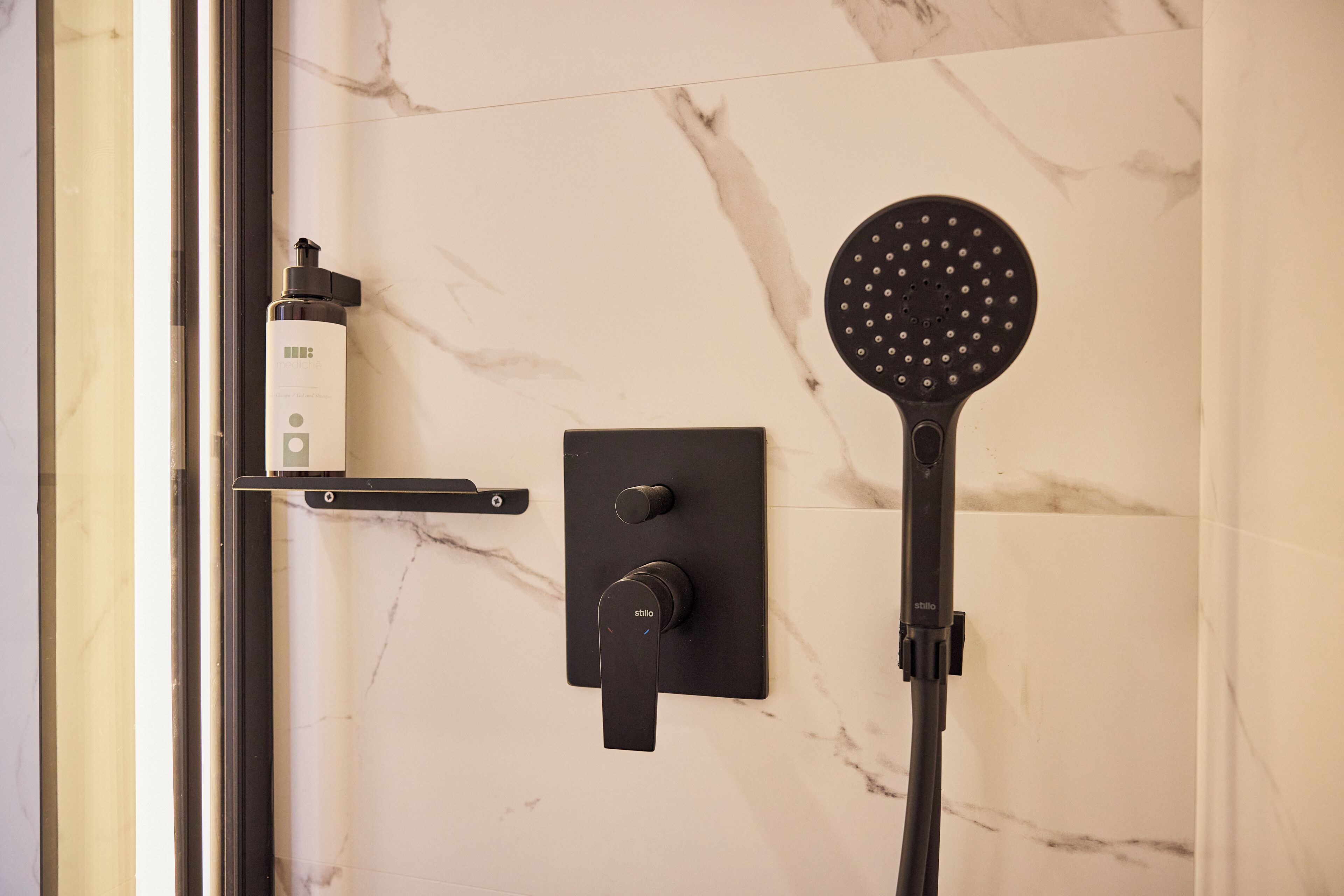 A modern bathroom with a marble wall, a handheld showerhead, and a bottle of shampoo.