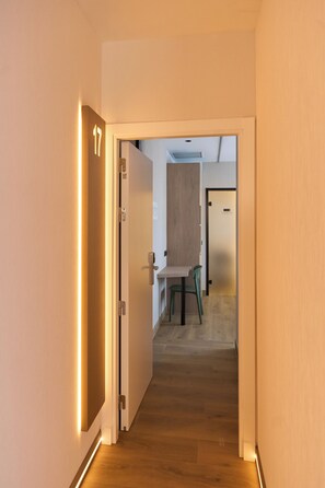 A hallway with a wooden floor, a door with a number, and a view into a room with a table and chairs.