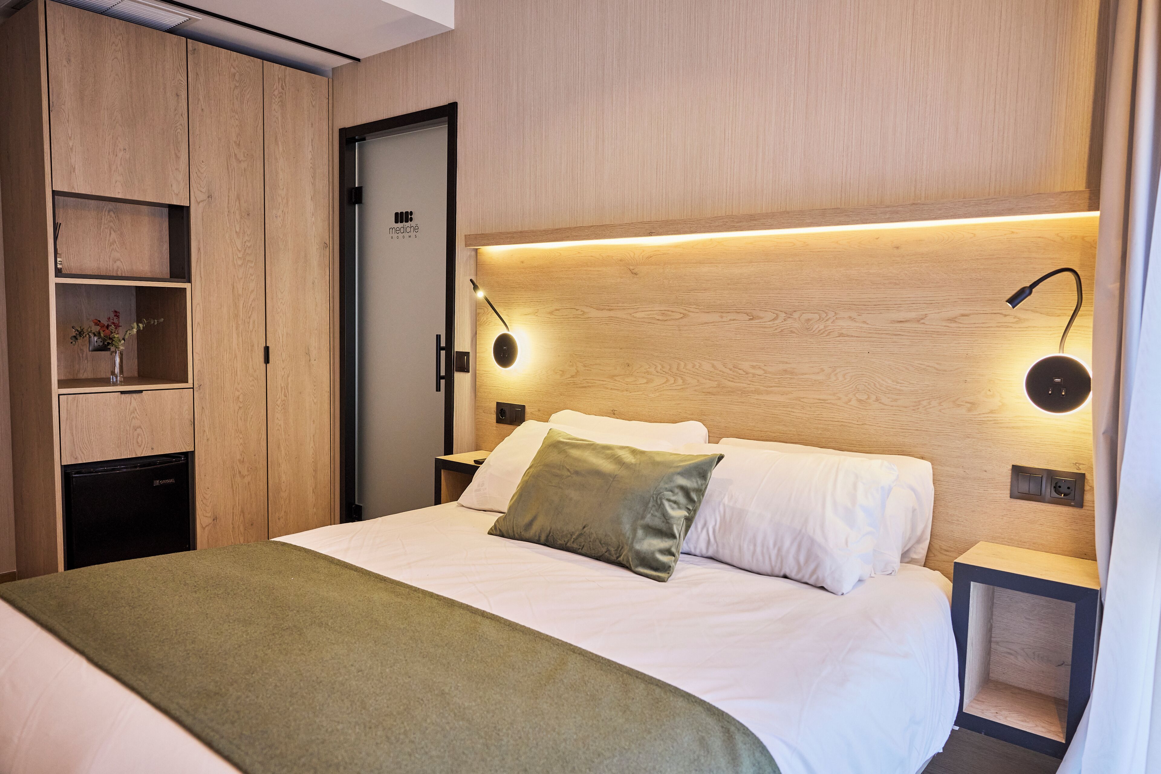Una habitación de hotel moderna con cabecera de madera, cama con ropa de cama blanca, una manta verde, una mesita de noche con una lámpara y un estante empotrado con un jarrón.