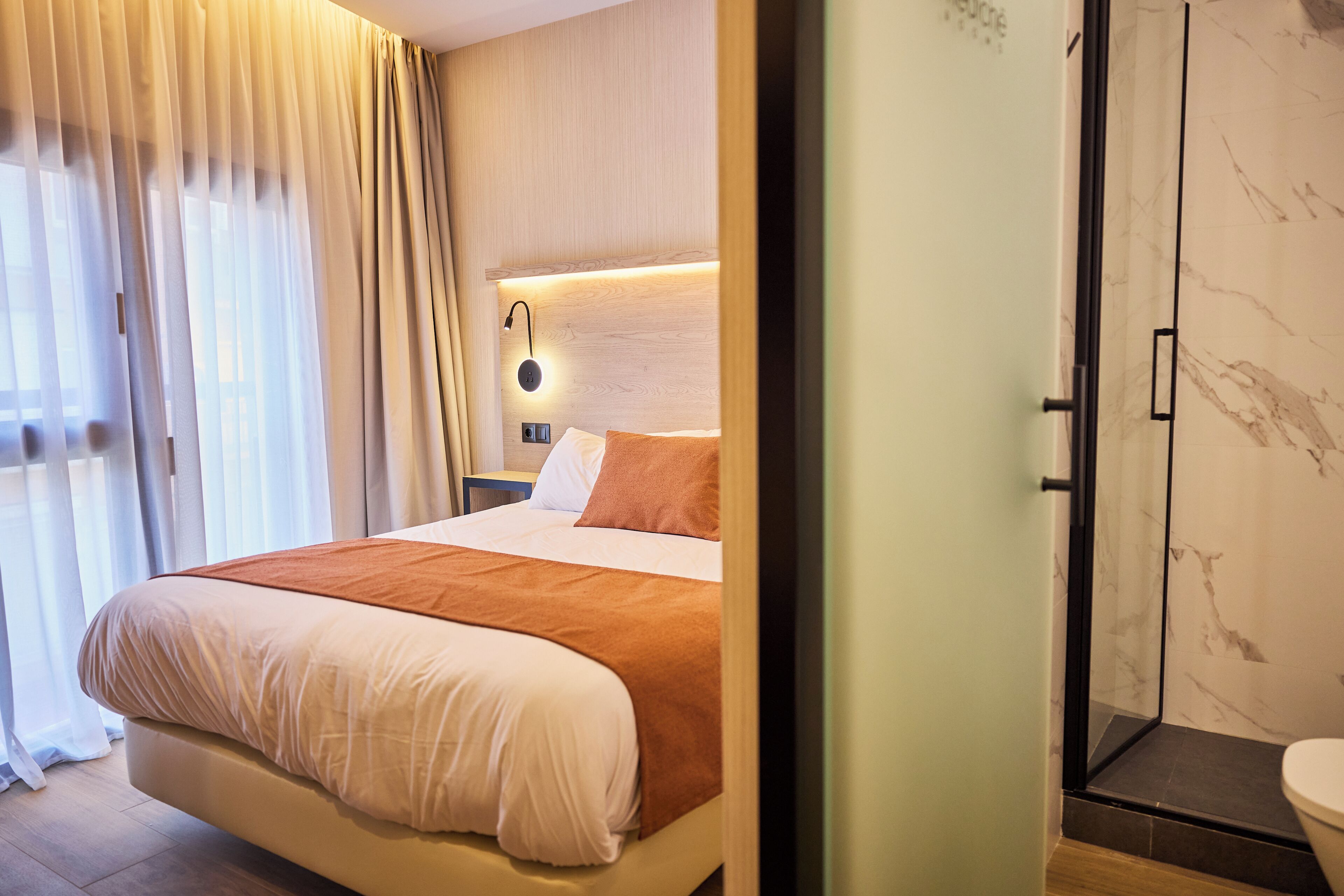 A hotel room with a bed, a nightstand, a wall-mounted lamp, and a shower area.