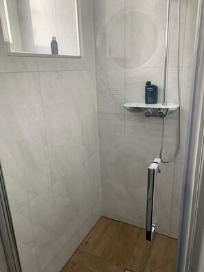 Une douche équipée d’un pommeau de douche à main, d’une étagère fixée au mur avec une bouteille, et d’une fenêtre située au-dessus de la douche.