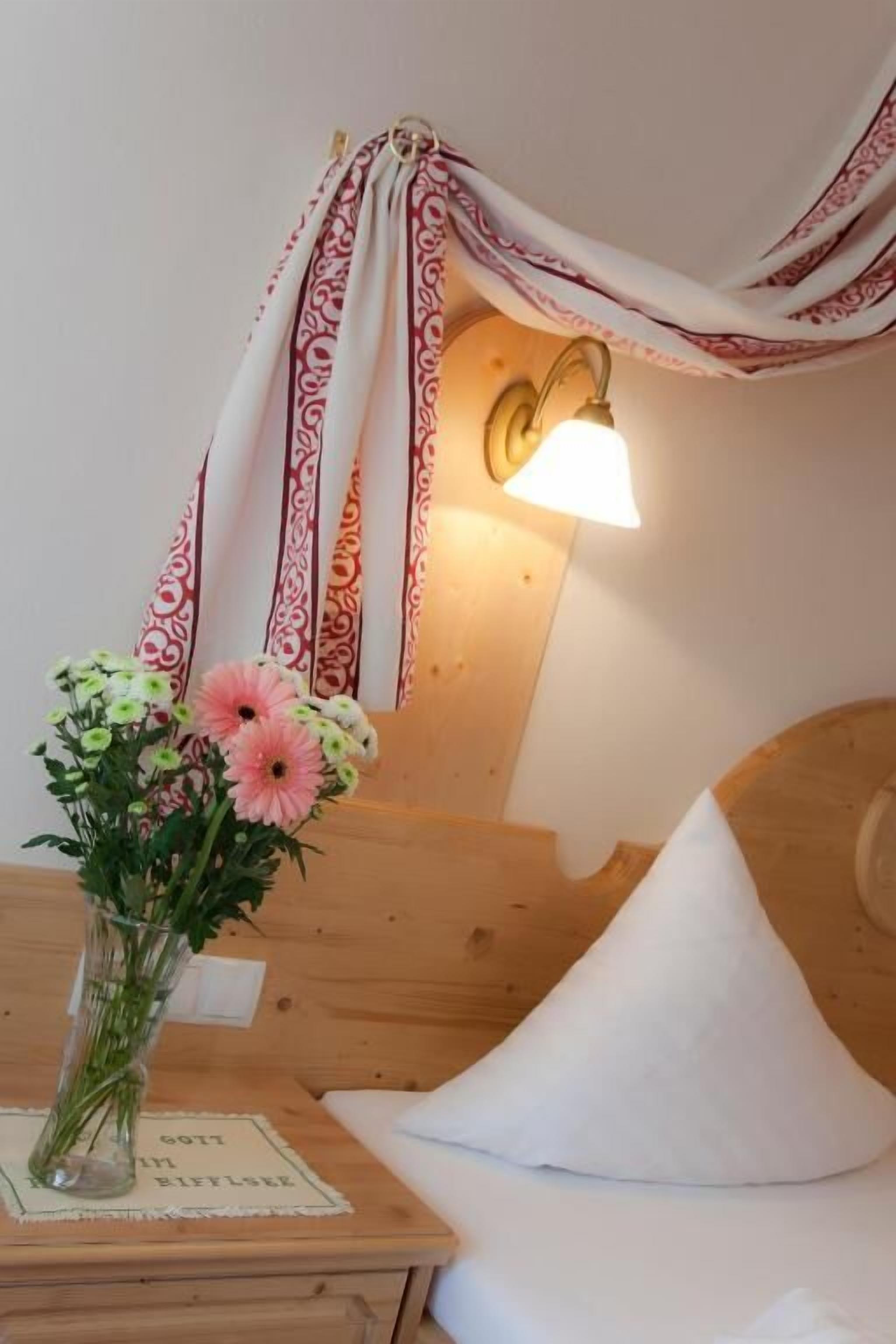 Una cama de madera con un almohadón blanco, una mesita de noche con un florero y una lámpara fijada a la pared.