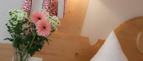 Una cama de madera con un almohadón blanco, una mesita de noche con un florero y una lámpara fijada a la pared.