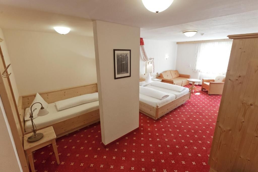 A hotel room with a red carpet, two beds, a wooden bench, a desk with a lamp, and a framed picture on the wall.