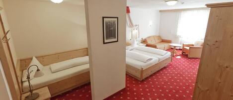 A hotel room with a red carpet, two beds, a wooden bench, a desk with a lamp, and a framed picture on the wall.
