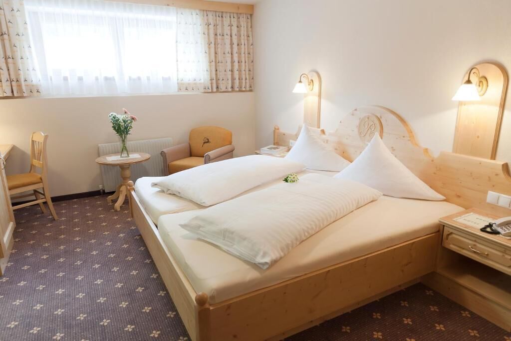 A hotel room with a bed, bedside tables, a chair, a small table with flowers, and a window with curtains.