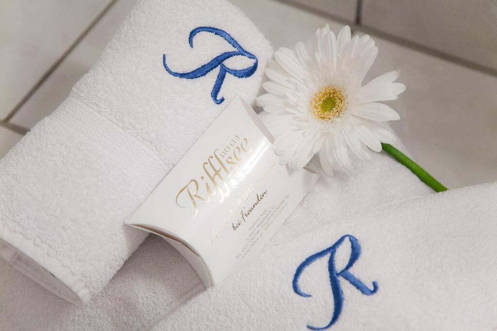 Una toalla blanca con letras bordadas y una etiqueta con nombre, una flor blanca, y una etiqueta con texto.