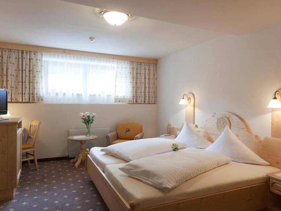 A hotel room with a bed, a desk with a computer, a chair, a small table with flowers, and a window with curtains.