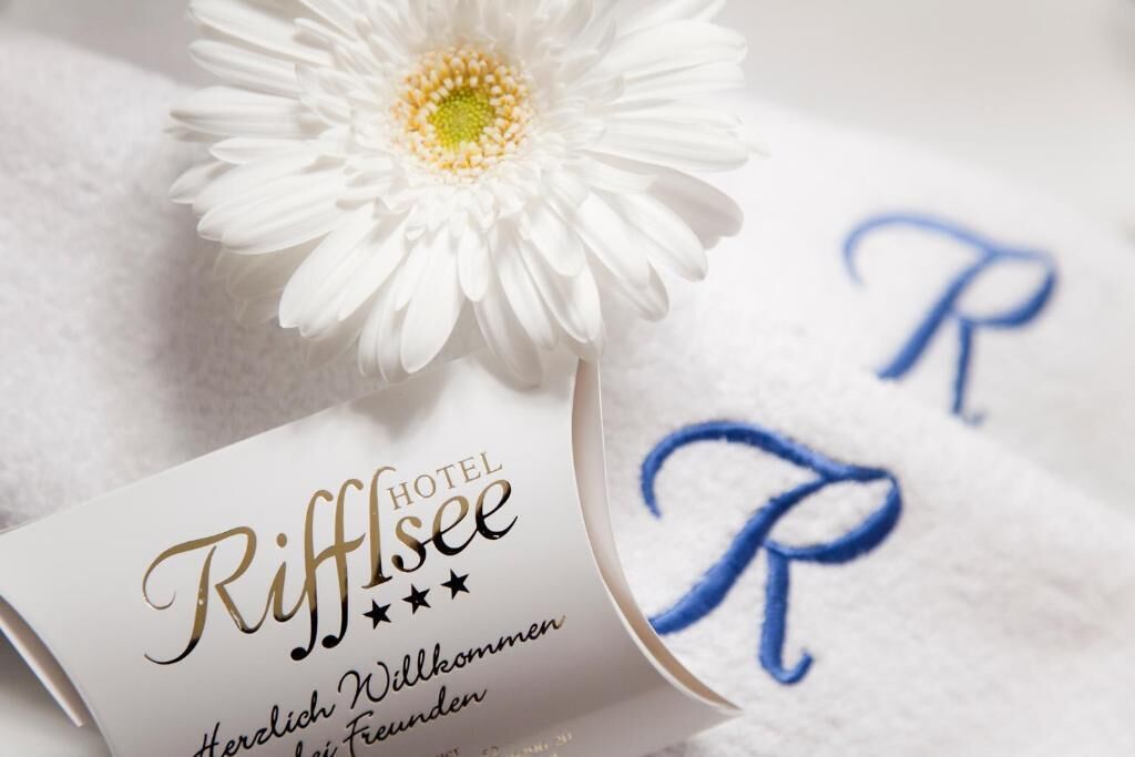 A hotel room with a white bedspread, a white flower, and a card with 'Riffisee' and 'Herzlich Willkommen' text.