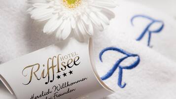 Una habitación de hotel con una colcha blanca, una flor blanca y una tarjeta con la leyenda 'Riffisee' y 'Herzlich Willkommen'.