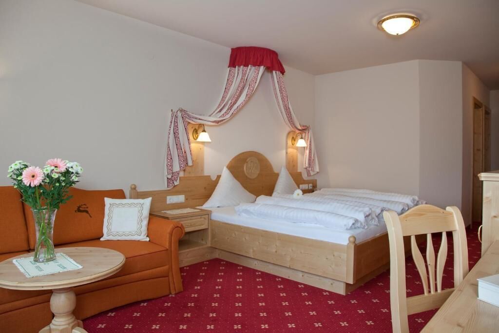 A bedroom with a bed, a wooden nightstand, a chair, a small table with a vase of flowers, and a red patterned carpet.