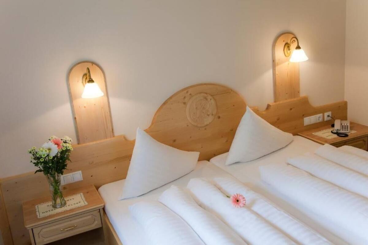 Une chambre d’hôtel avec une tête de lit en bois, deux lampes de chevet, une table de chevet ornée d’un vase de fleurs et un lit recouvert de draps blancs.